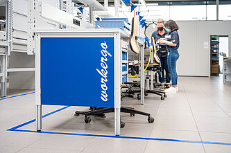 Ansicht des ergonimischen Arbeitsplatzes. Im Hintergrund stehen zwei Mitarbeiterinnen.